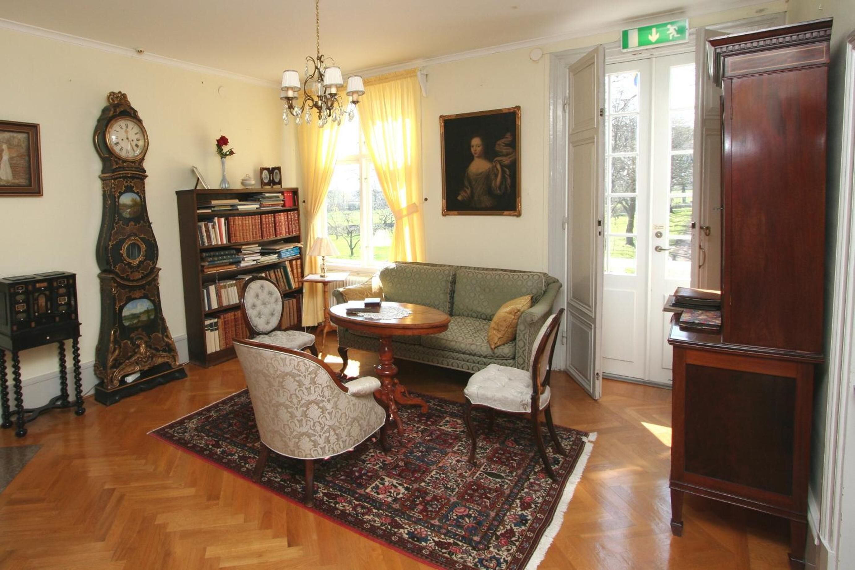 A traditional room with a wooden floor, a green patterned sofa, a round wooden table, a large wooden cabinet, a tall grandfather clock, and a bookshelf filled with books.