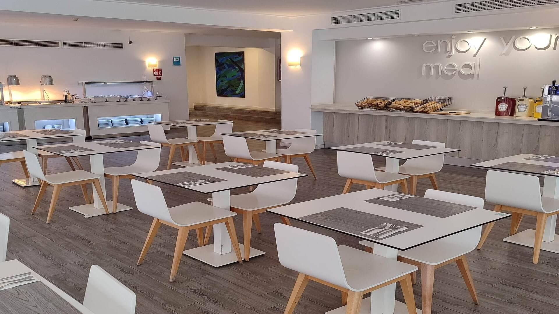A modern dining area with white tables and chairs, a counter with food items, and a wall with the text "enjoy your meal".