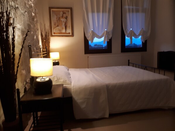 A hotel room with a bed, a desk with a lamp, a radiator, and a painting on the wall.