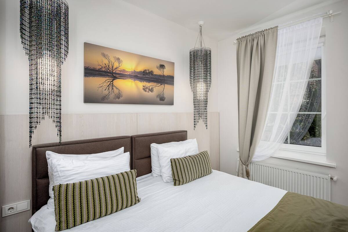 Habitación de hotel con una cama grande, un cuadro de un atardecer, una araña y una ventana con cortinas.