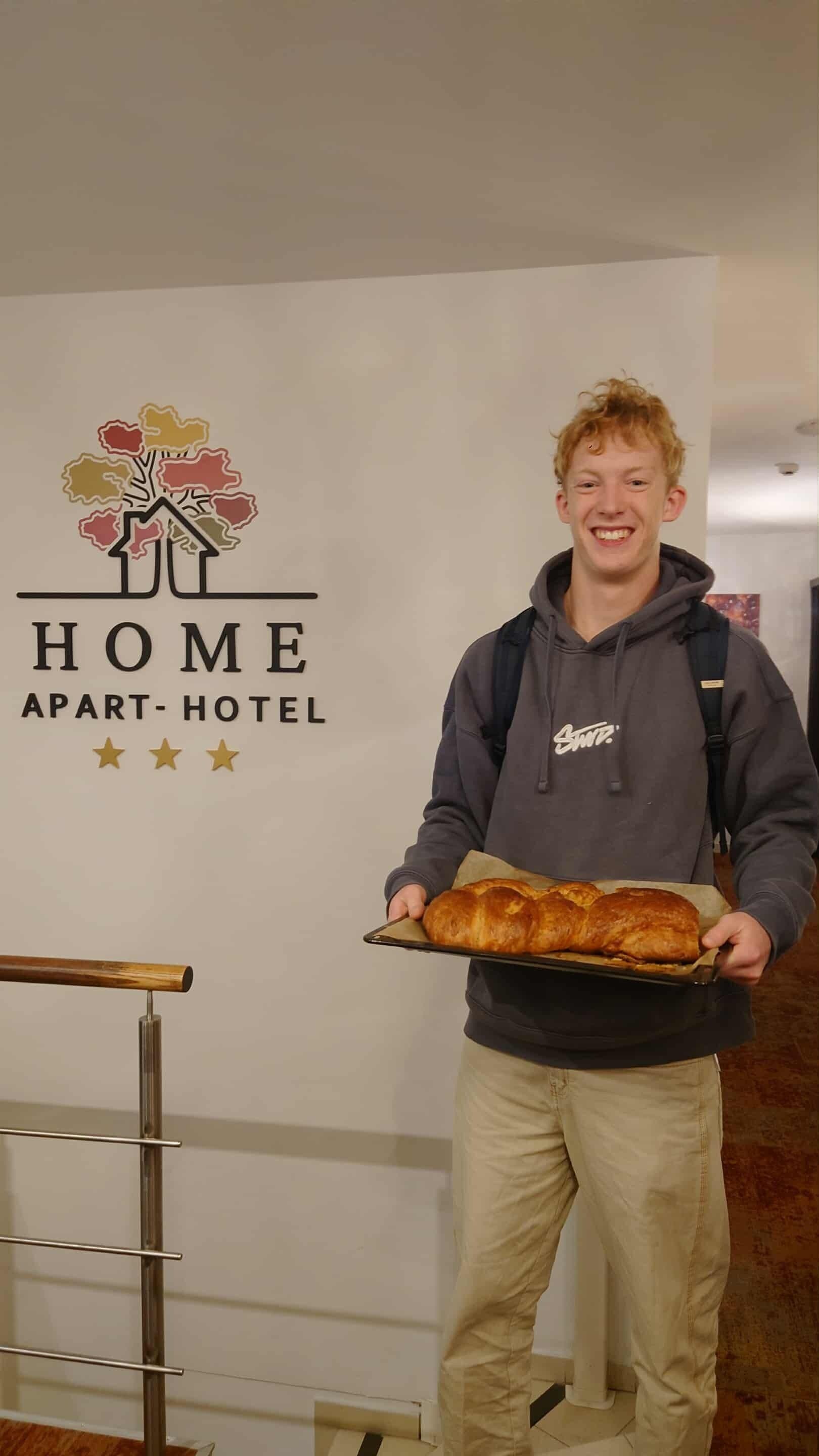 Una persona sostiene un pan frente a un letrero que dice 'HOME APART-HOTEL' con tres estrellas.