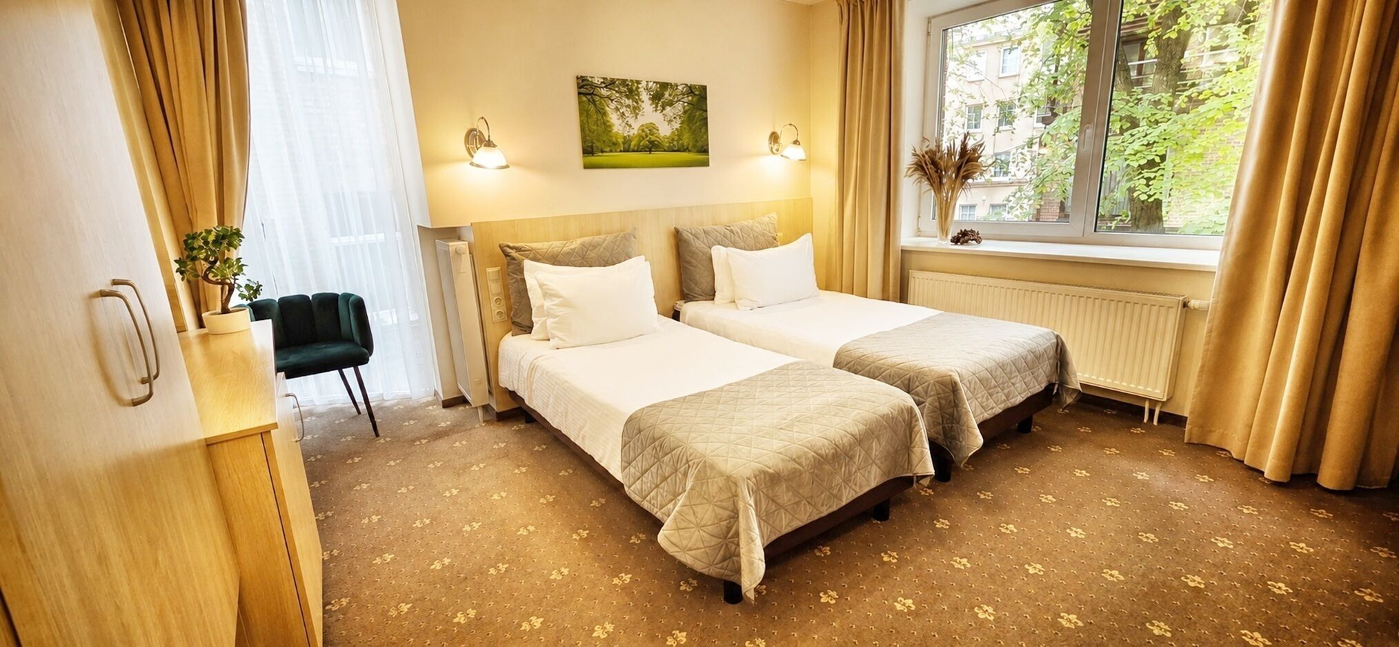 A hotel room with two beds, a desk, a chair, a window with a view of trees, and a radiator.