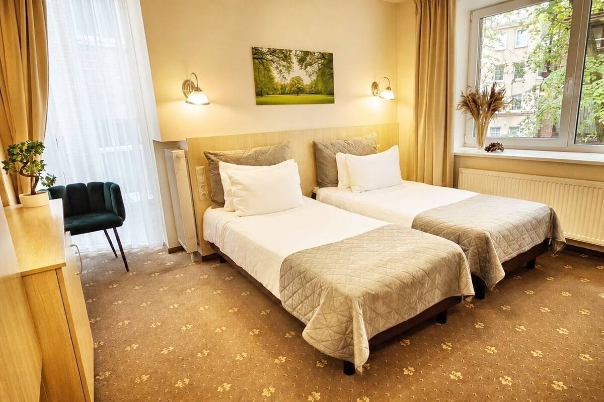 Habitación de hotel con dos camas, un escritorio, una silla, un ventanal con vistas a árboles y un radiador.
