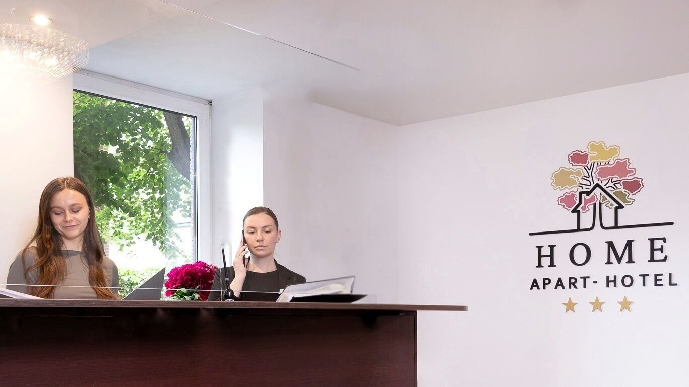 A reception desk with two staff members, a window with a view of trees, and a wall with the logo 'HOME APART-HOTEL' and three stars.