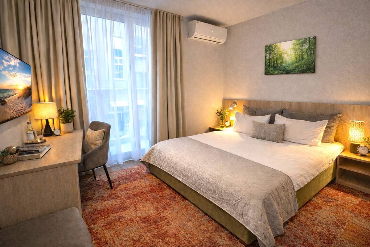 Una habitación de hotel con una cama grande, un escritorio con lámpara, una silla, un cuadro en la pared y un equipo de aire acondicionado.