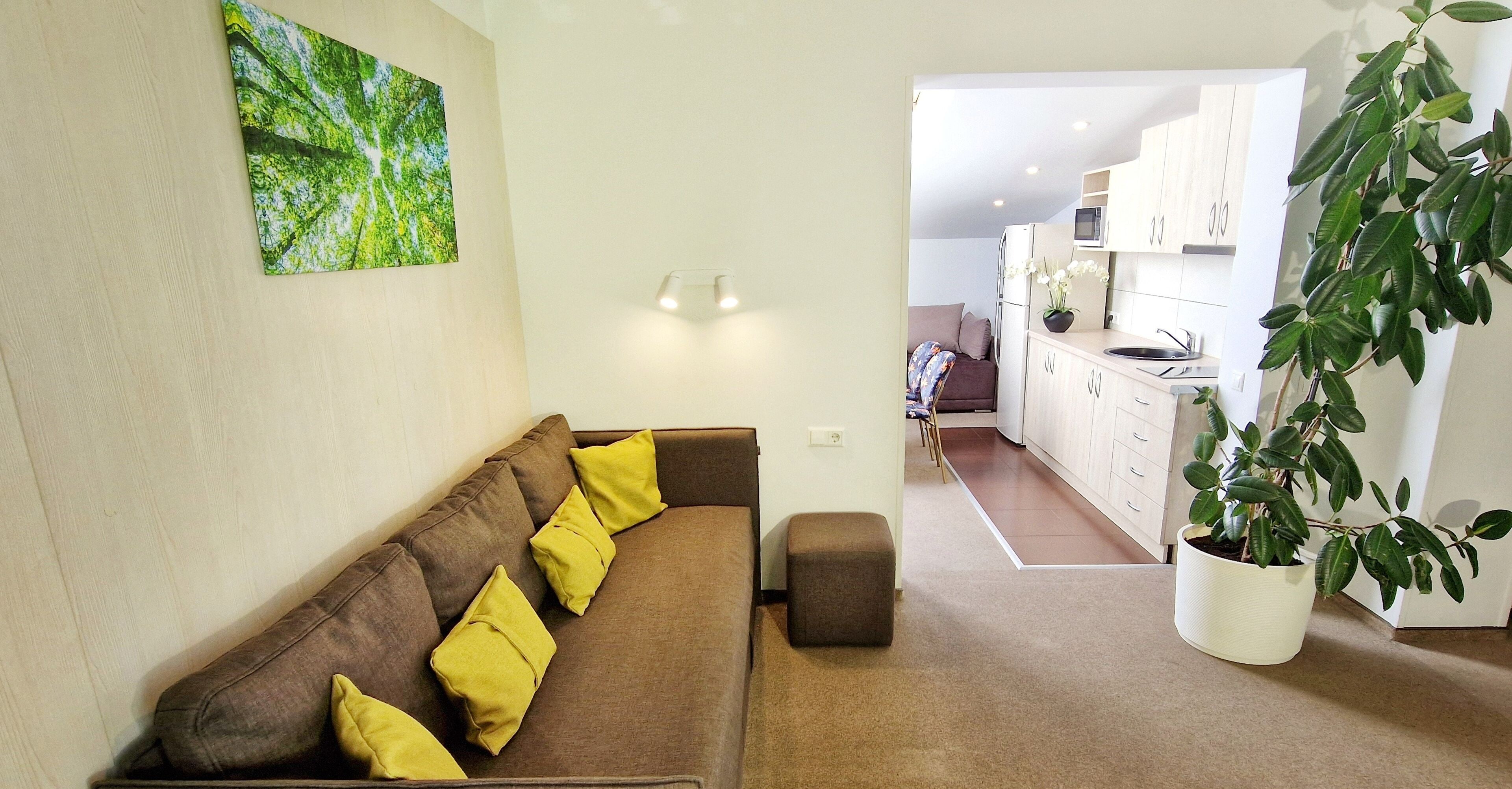A living room with a brown sofa, yellow cushions, a small ottoman, a potted plant, and a painting on the wall.