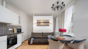 A modern kitchen with white cabinets, a black countertop, and a tiled backsplash. There is a dining area with a round table, two chairs, and a vase with flowers. A sofa is placed in the center of the room, and a painting hangs on the wall. - HOME Apart-Hotel Vilnius (Vilnius)
