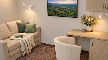 A cozy living area with a sofa, a small desk, and a chair. There is a framed landscape picture on the wall and a vase with flowers on the desk.