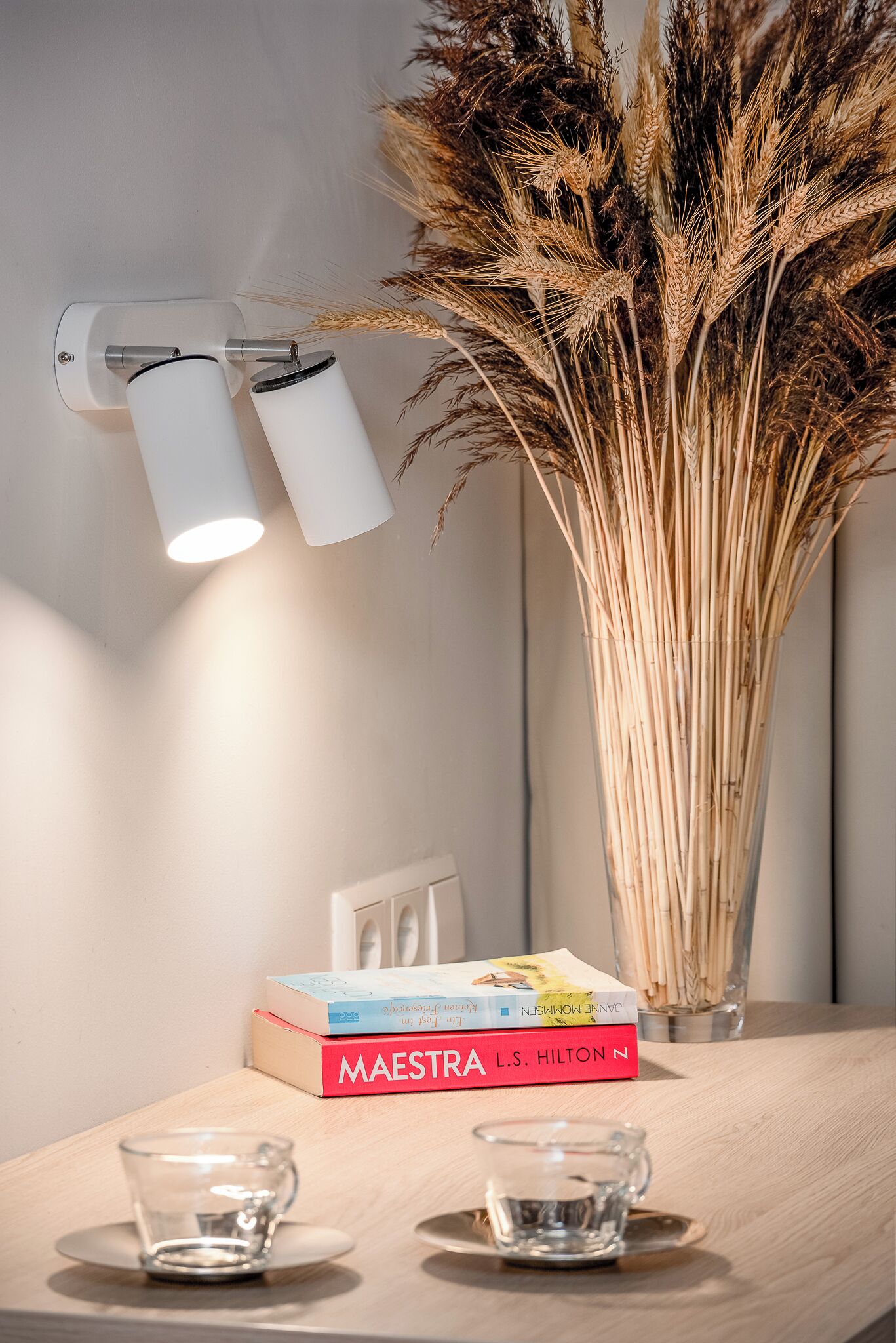 A table with a clear glass cup, a stack of books, and a vase with dried plants.