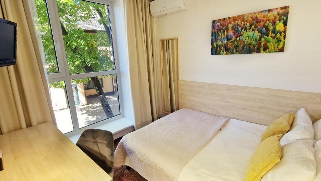A hotel room with a bed, a desk, a chair, a window with curtains, and a painting on the wall.