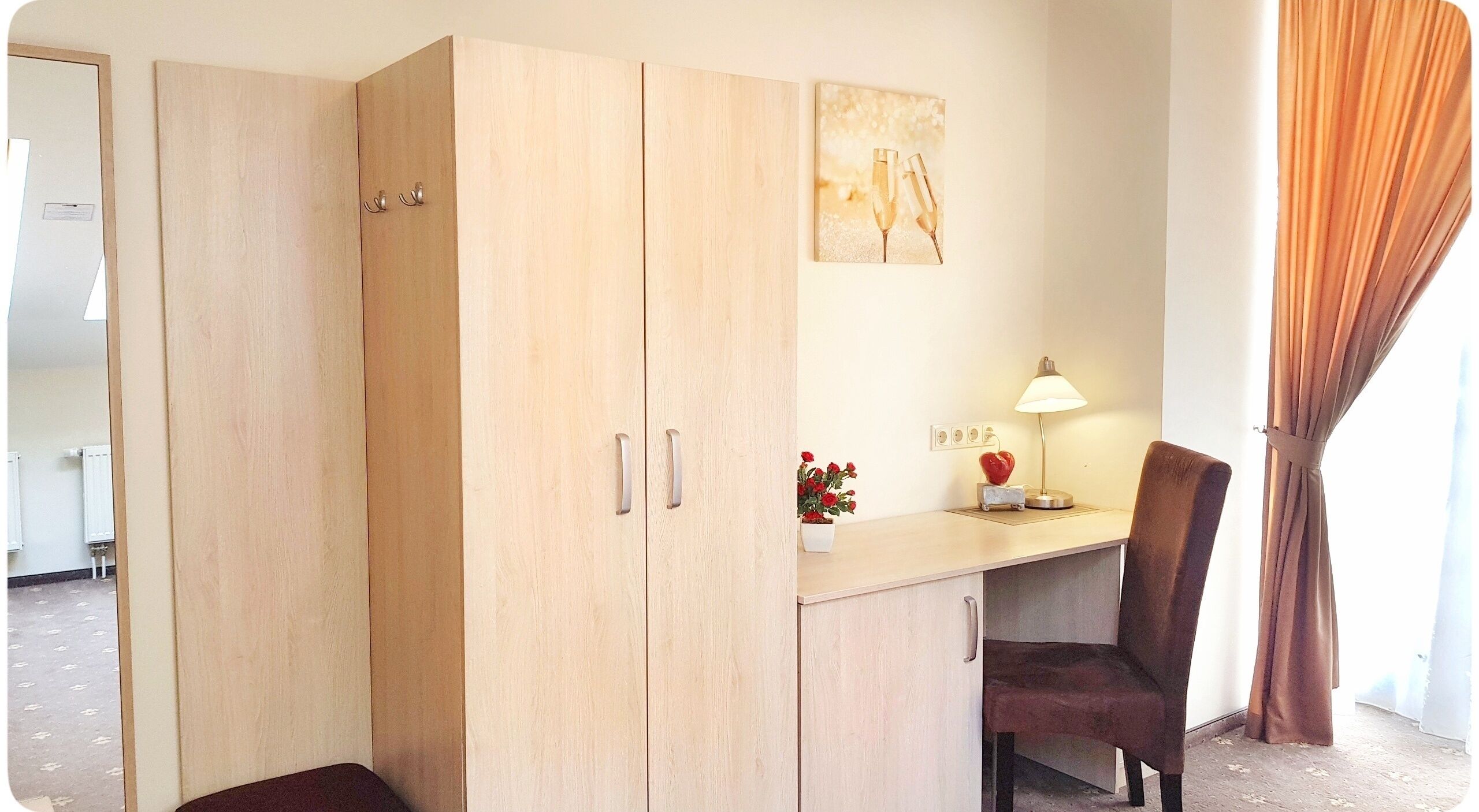 A room with a wooden wardrobe, a desk with a lamp, a chair, and a small potted plant.