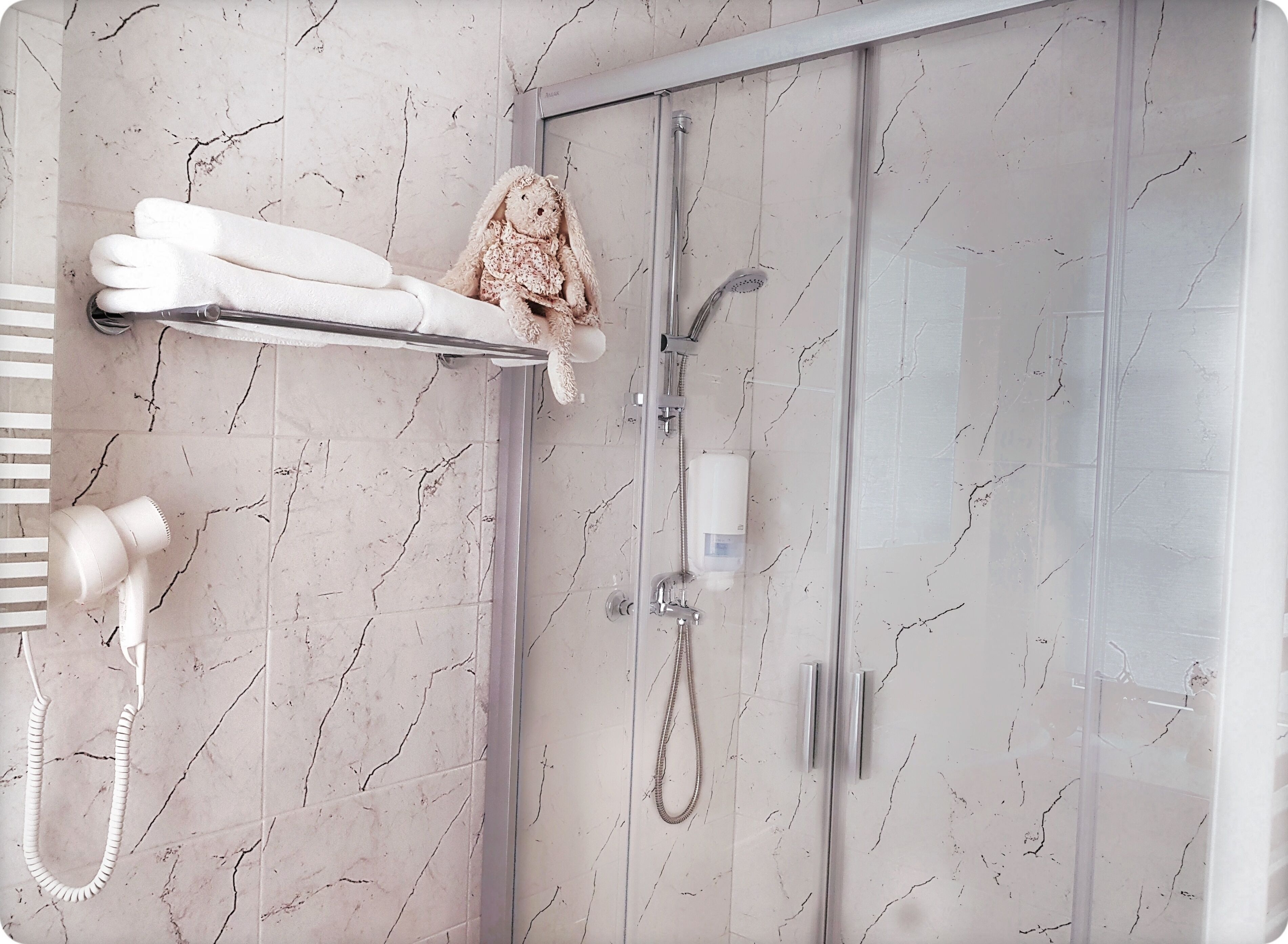 Une salle de bain avec un sol en marbre, une cabine de douche en verre, un porte-serviettes orné d’un jouet en peluche et une douchette à main.