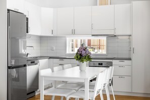 A modern kitchen with white cabinetry, a stainless steel refrigerator, a dining table with white chairs, and a vase with flowers on the table.