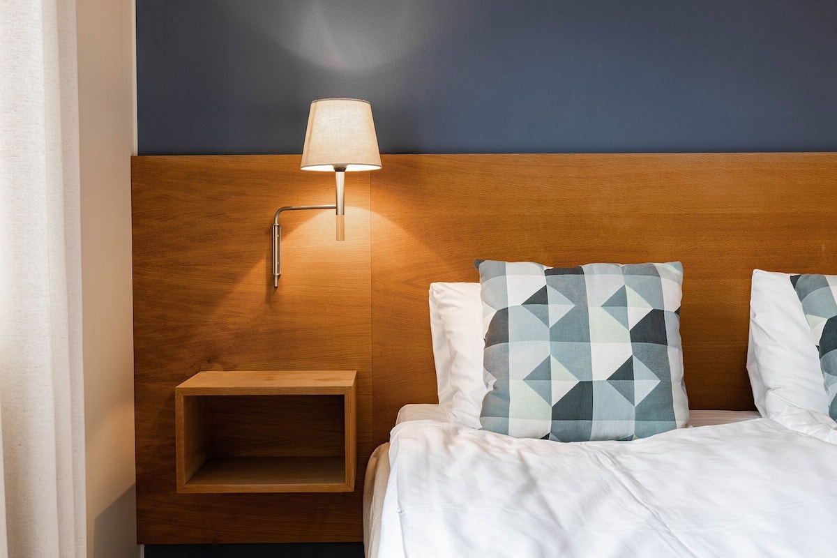 Ein Hotelzimmer mit einem Holzkopfstück, einer Nachttischlampe, einem Nachttisch und gemusterten Kissen.