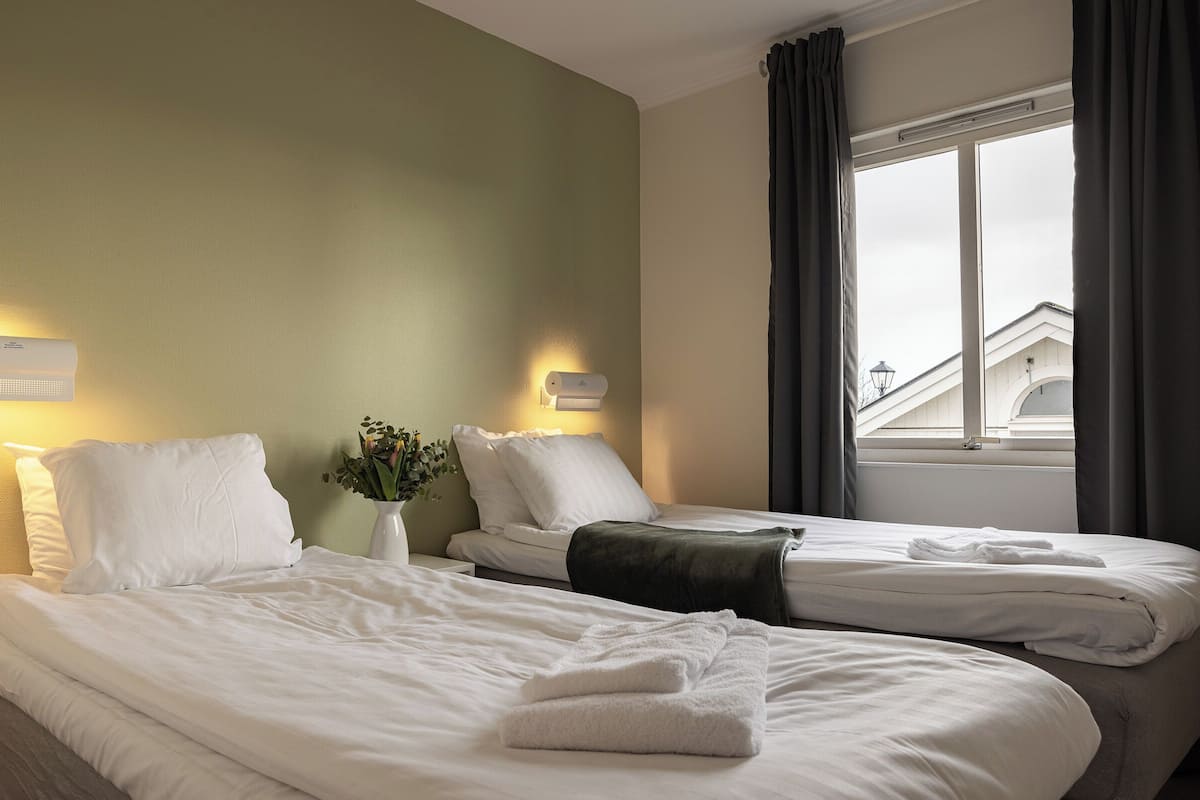 Ein Hotelzimmer mit zwei Betten, einem Fenster mit Vorhängen, einer Wandleuchte und einer Vase mit Blumen.