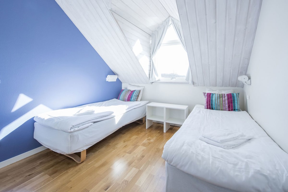 Ein Zimmer mit schrägem Dach, zwei Betten, einem kleinen Tisch und einem Fenster.