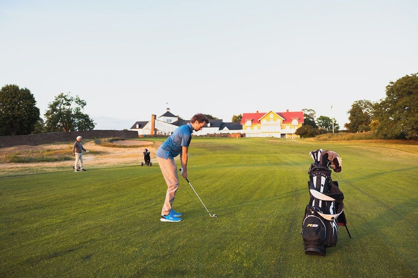 A golf course with a player preparing to putt, a golf bag, and a house in the background.