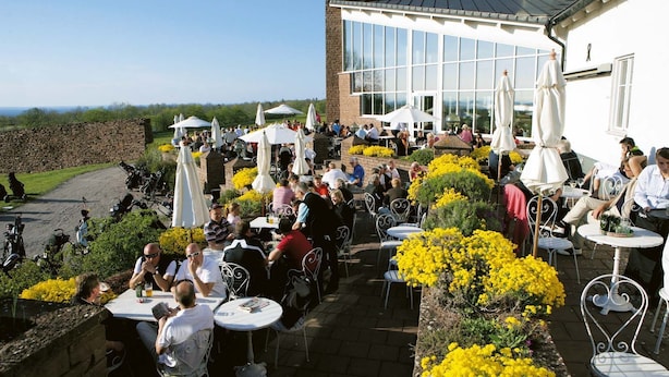 Un espace extérieur aménagé en terrasse, avec des tables et des chaises, entouré de fleurs jaunes et d’un mur en pierre.