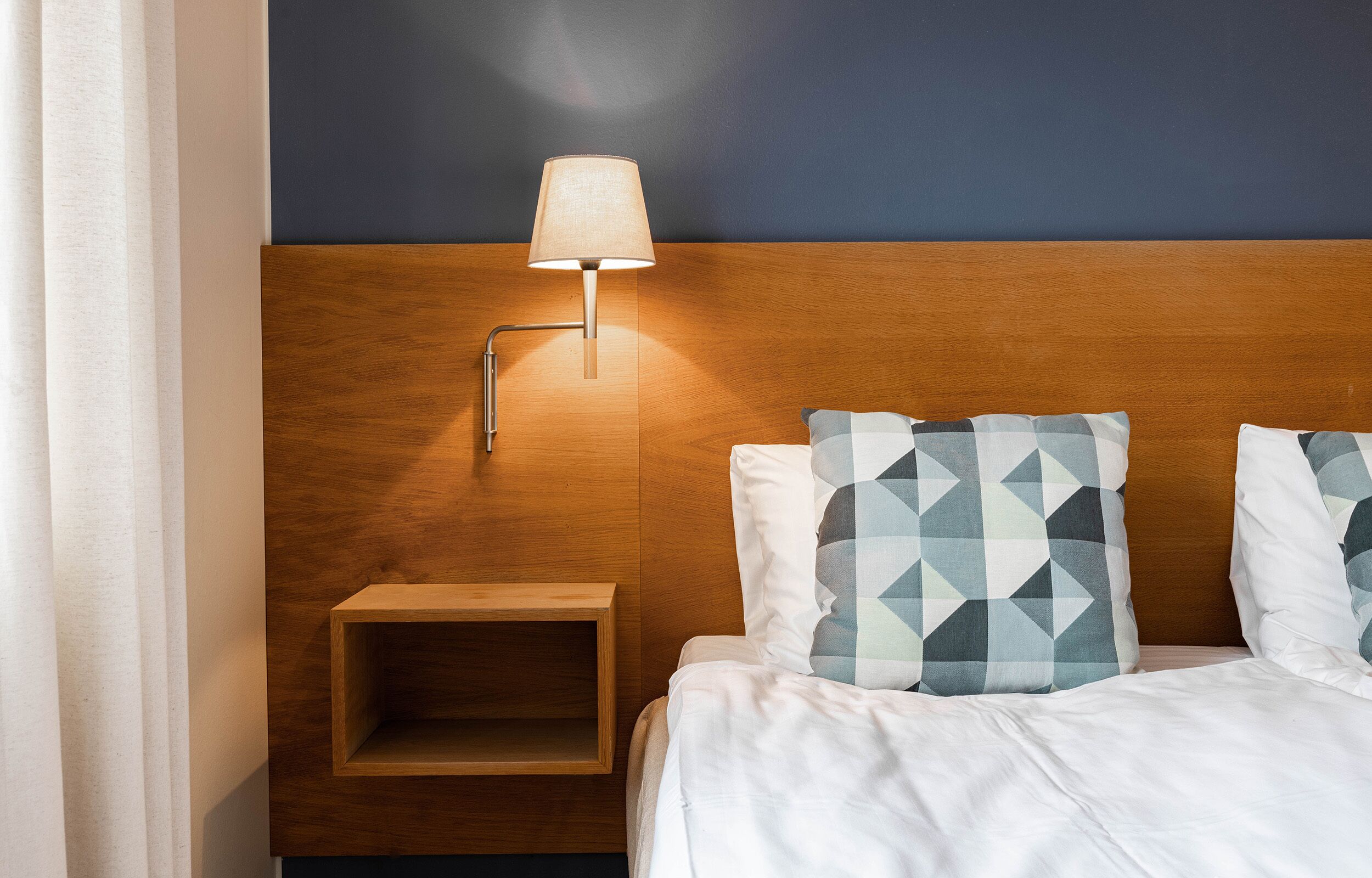 Camera d'albergo con testiera in legno, comodino e lampada. Due cuscini con motivo geometrico e biancheria da letto bianca.