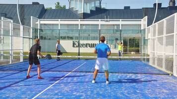 En indendørs tennisbane med net, et glasvinduer og en bygning med skiltning, der viser 'Ekerum' i baggrunden.