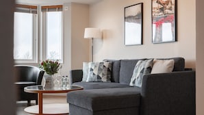 Un salon moderne avec un canapé gris, une table basse ronde et un fauteuil noir. Deux tableaux encadrés ornent le mur, et une lampe sur pied est placée dans un coin.