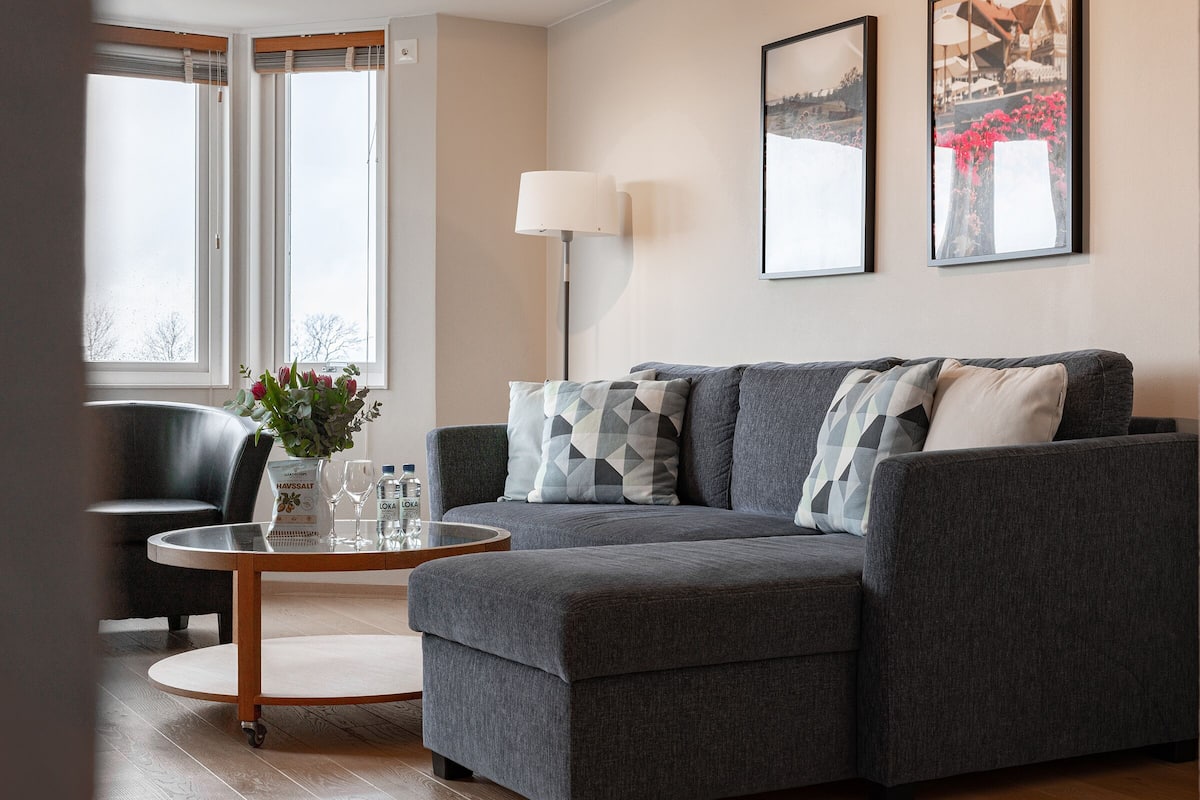 Ein modernes Wohnzimmer mit einem grauen Sofa, einem runden Couchtisch und einem schwarzen Sessel. An der Wand hängen zwei gerahmte Bilder und eine Stehlampe steht im Raum.