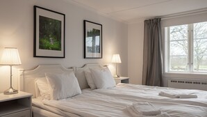 Une chambre d’hôtel avec un grand lit, deux tables de chevet avec des lampes, des tableaux encadrés au mur et une fenêtre avec des rideaux.