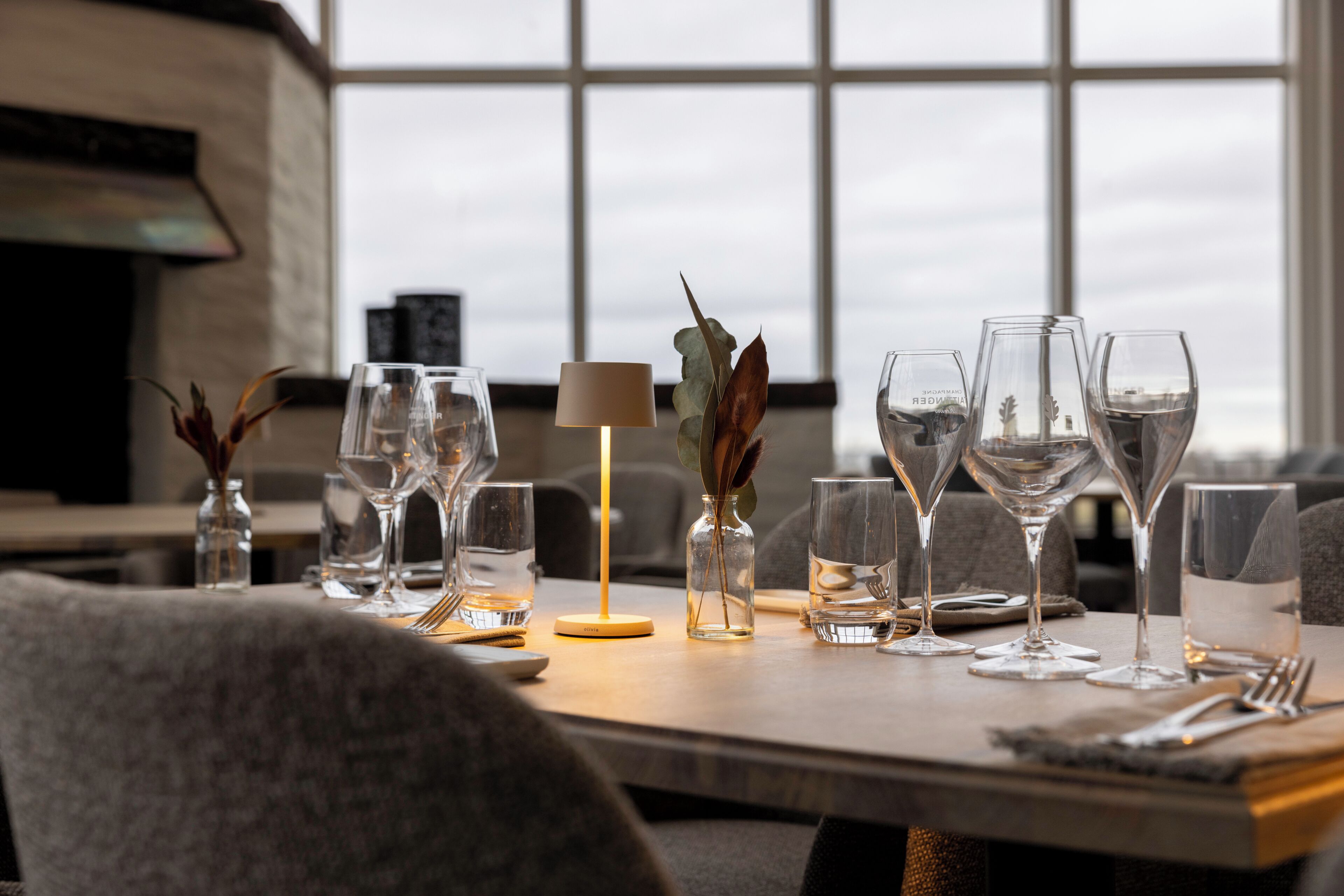Une table à manger dressée avec différents types de verres et une lampe, avec vue sur le ciel par une fenêtre.
