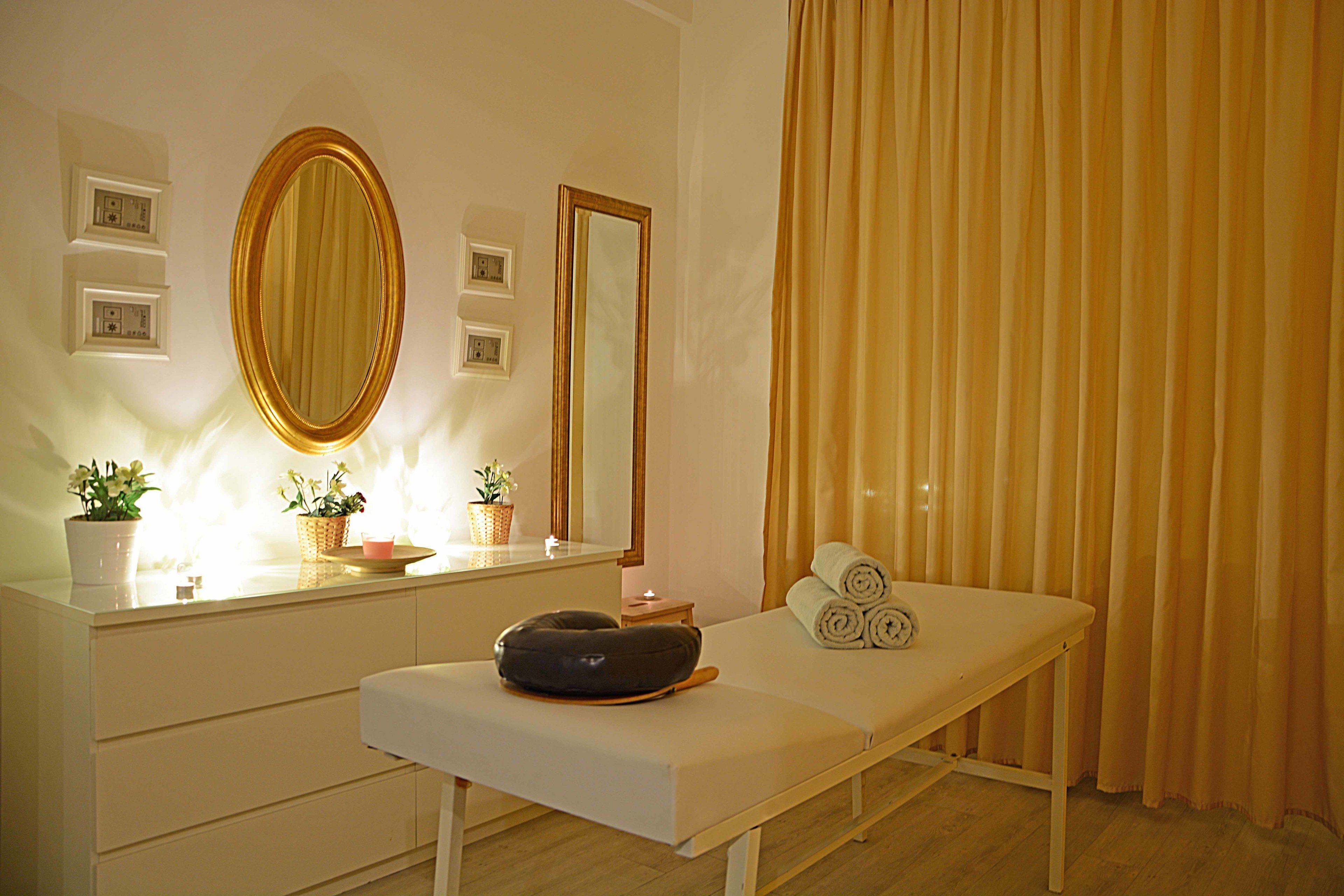 A room with a vanity, a mirror, a massage table, and a window with curtains.