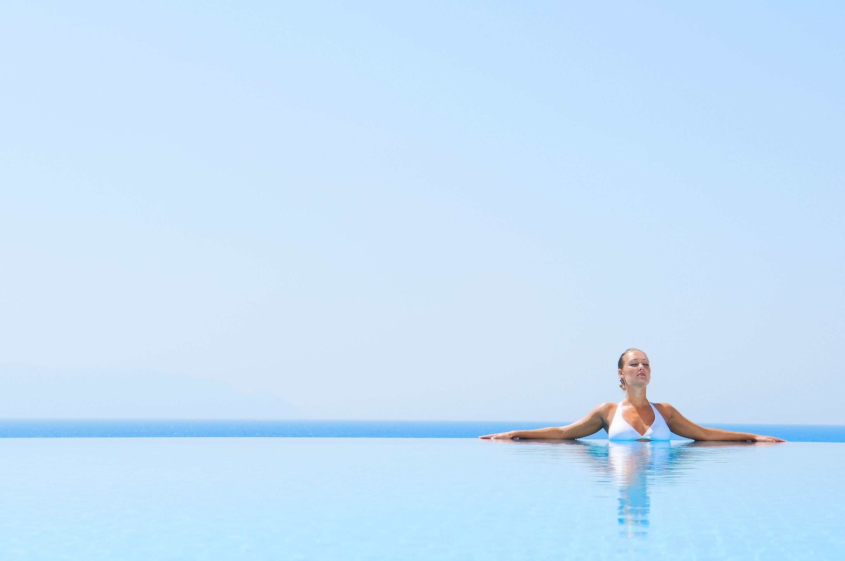 Une personne vêtue d’un maillot de bain blanc se détend dans une piscine à débordement aux eaux d’un bleu limpide, avec une vue sur l’horizon.