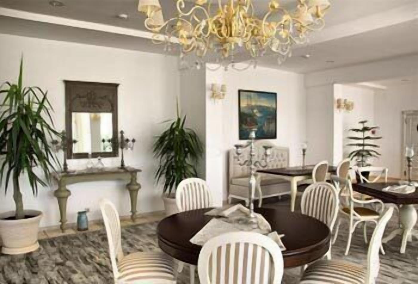 A dining area with a round table, white chairs, and a chandelier. There are potted plants and a painting on the walls.