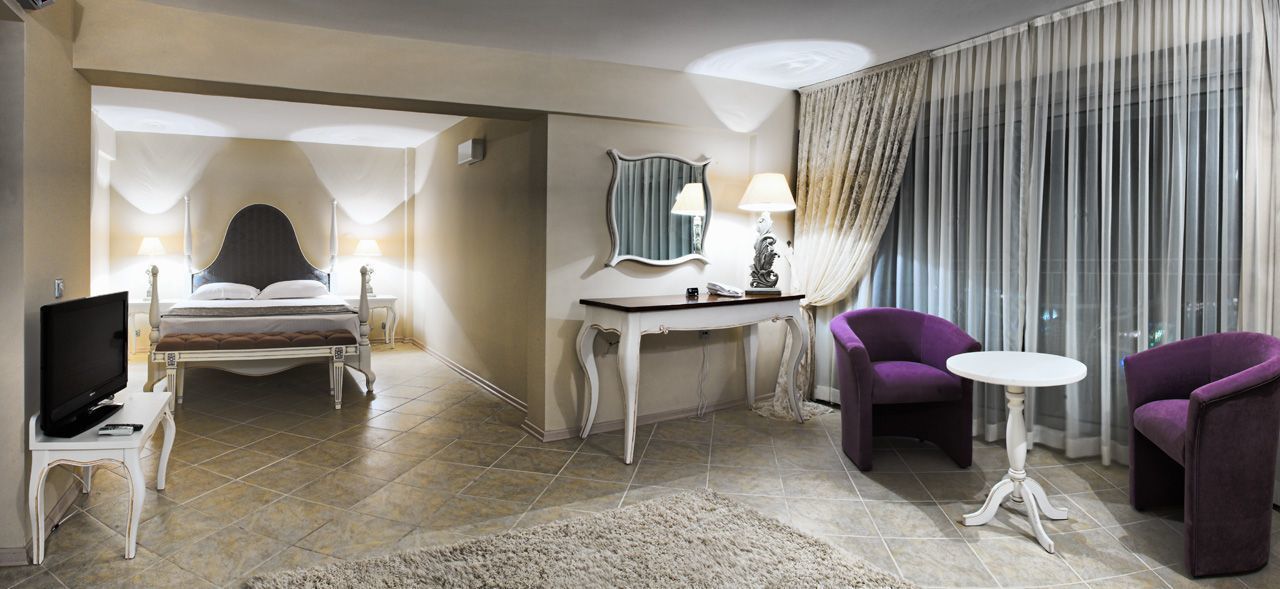 Una habitación de hotel con una cama, un escritorio con una lámpara, dos sillones morados y una pequeña mesa redonda.