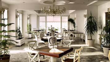A spacious dining area with a chandelier, white chairs, and potted plants.