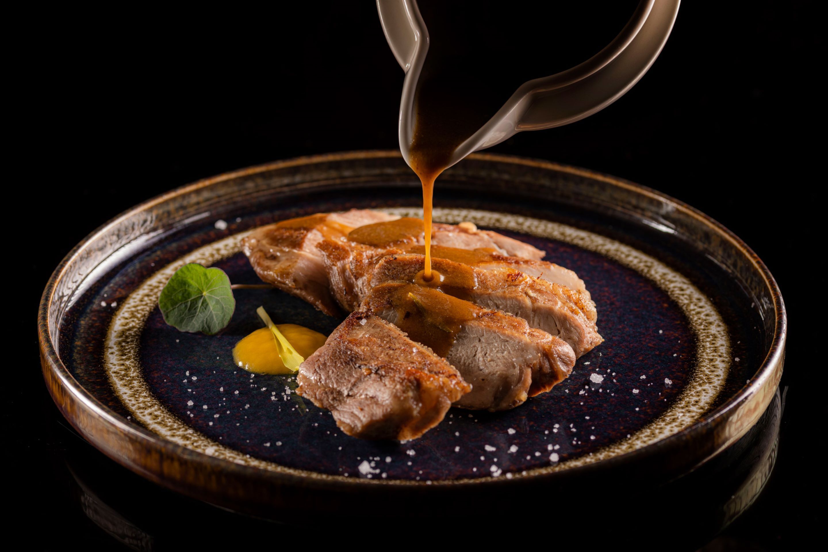 A plate with sliced meat and sauce, garnished with a leaf and a yellow item.