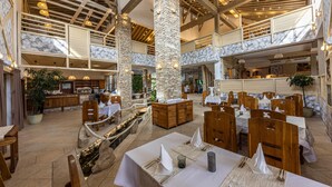 A spacious dining area with high ceilings, stone pillars, and wooden beams. Tables are set with white tablecloths and napkins.