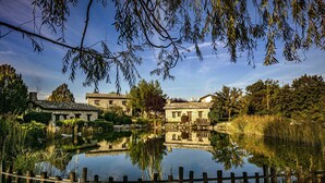A rustic stone building with a pond in front, surrounded by trees and a wooden fence.
