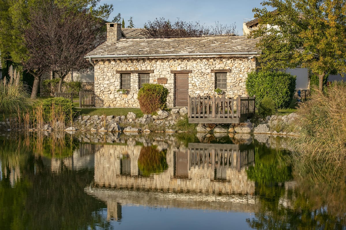A stone house with a pond in front, surrounded by trees and a wooden fence.