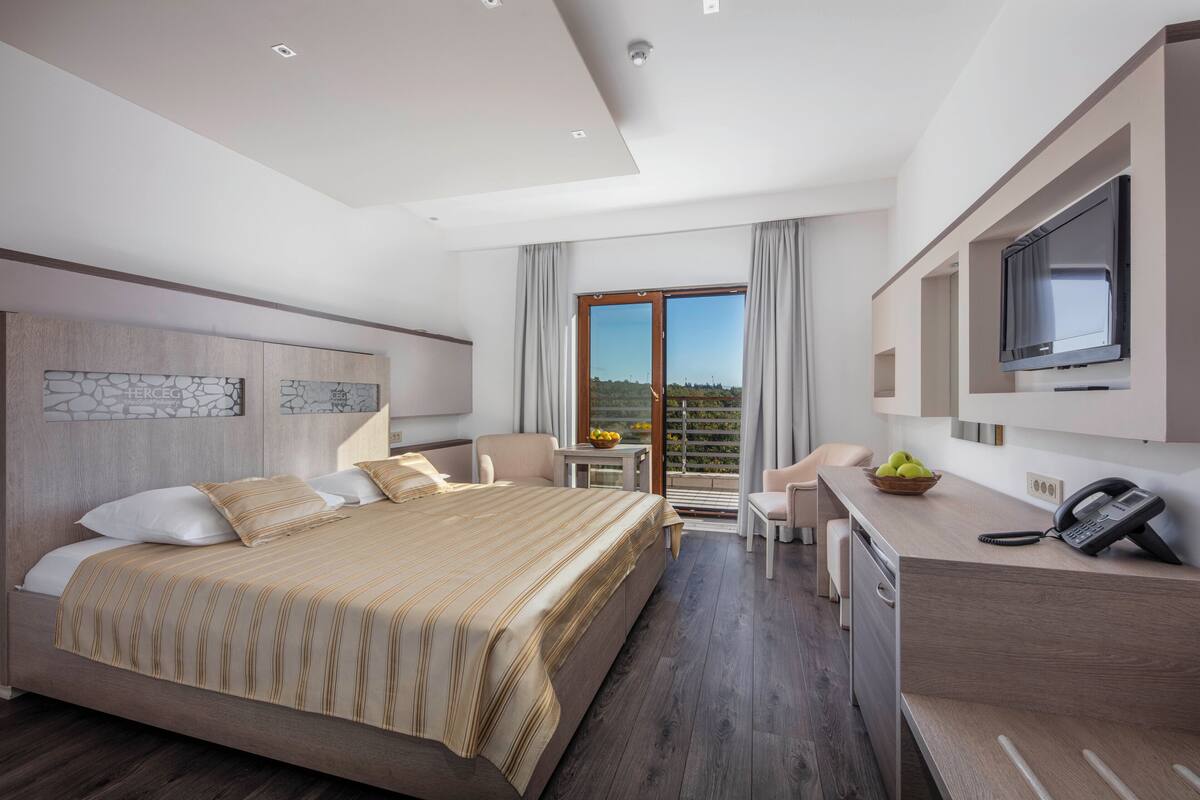 A modern hotel room with a large bed, a desk with a phone, a TV, and a balcony with a view.