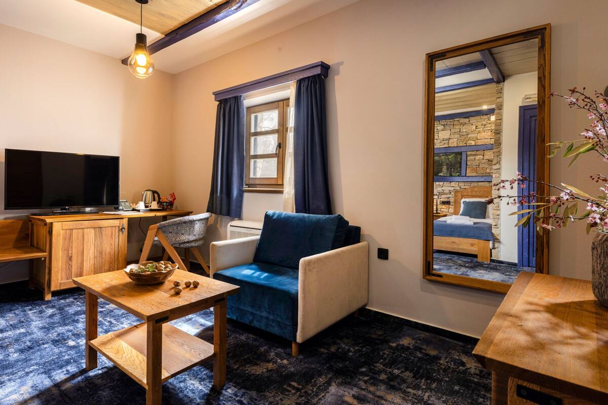 A cozy room with a wooden desk, a blue sofa, a television, and a large mirror reflecting a bedroom with a bed and window.