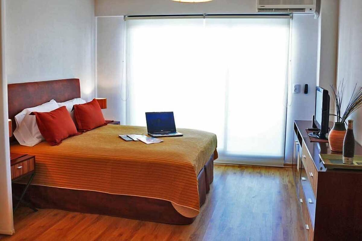 A bedroom with a bed, a laptop, a TV, and a desk with a vase.