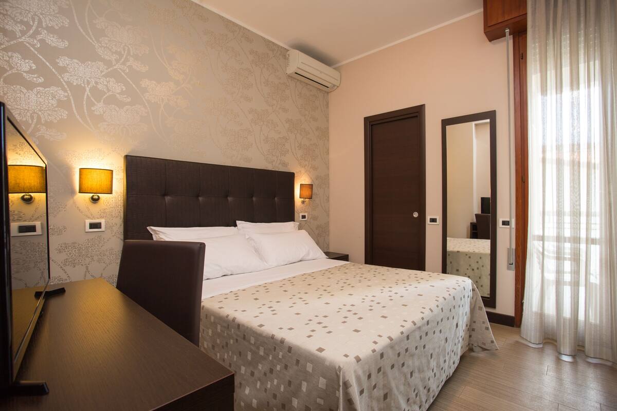 A hotel room with a bed, bedside lamps, a desk, a mirror, and an air conditioning unit.