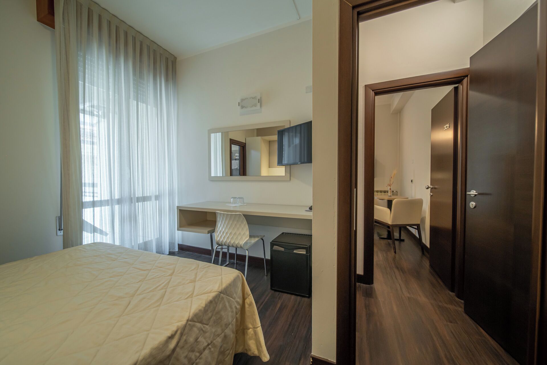 Een hotelkamer met een bed, een bureau, een stoel, een spiegel en een deur naar een andere kamer.