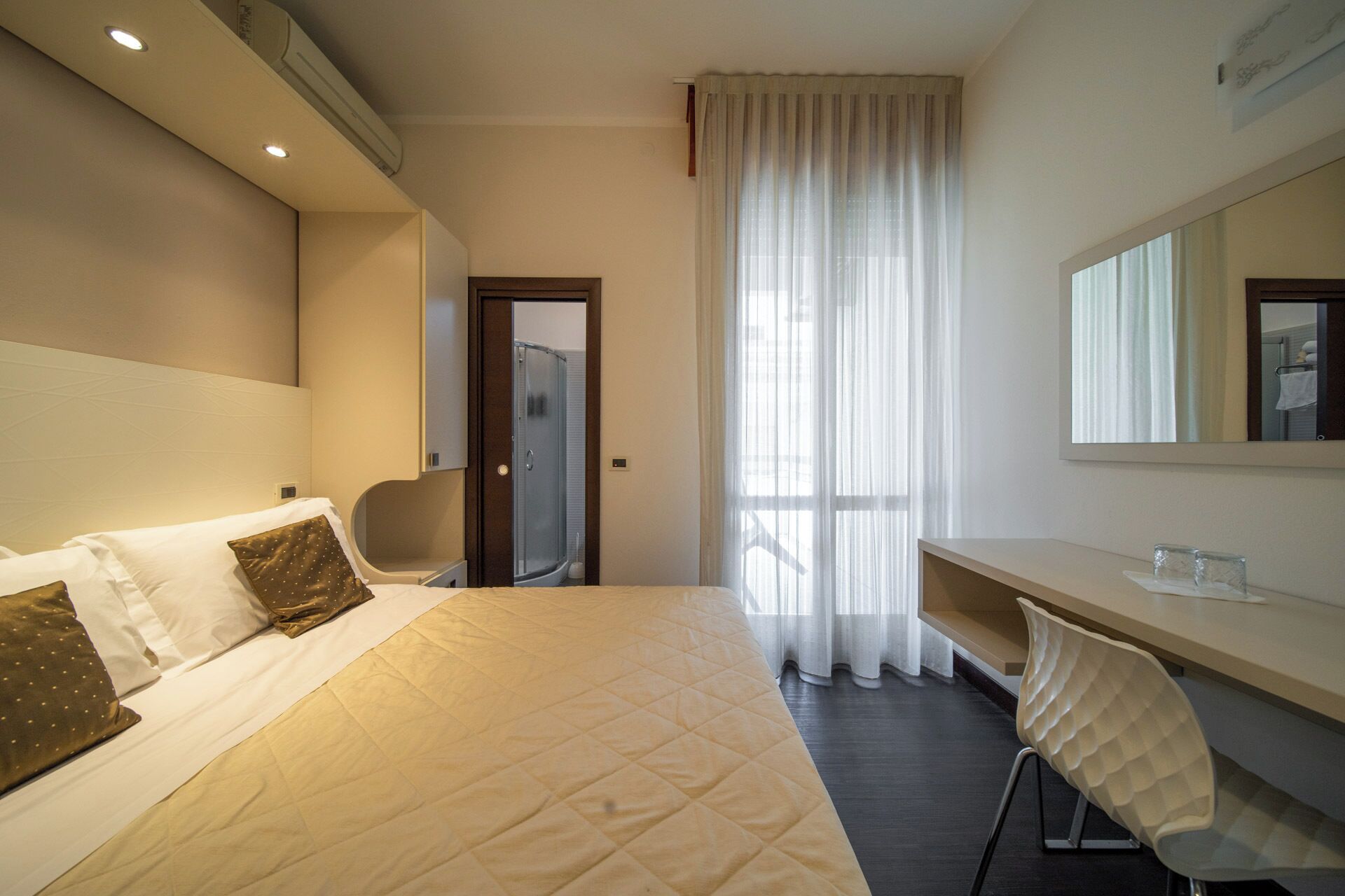 A hotel room with a bed, a desk, a chair, a shower, and a large window with curtains.