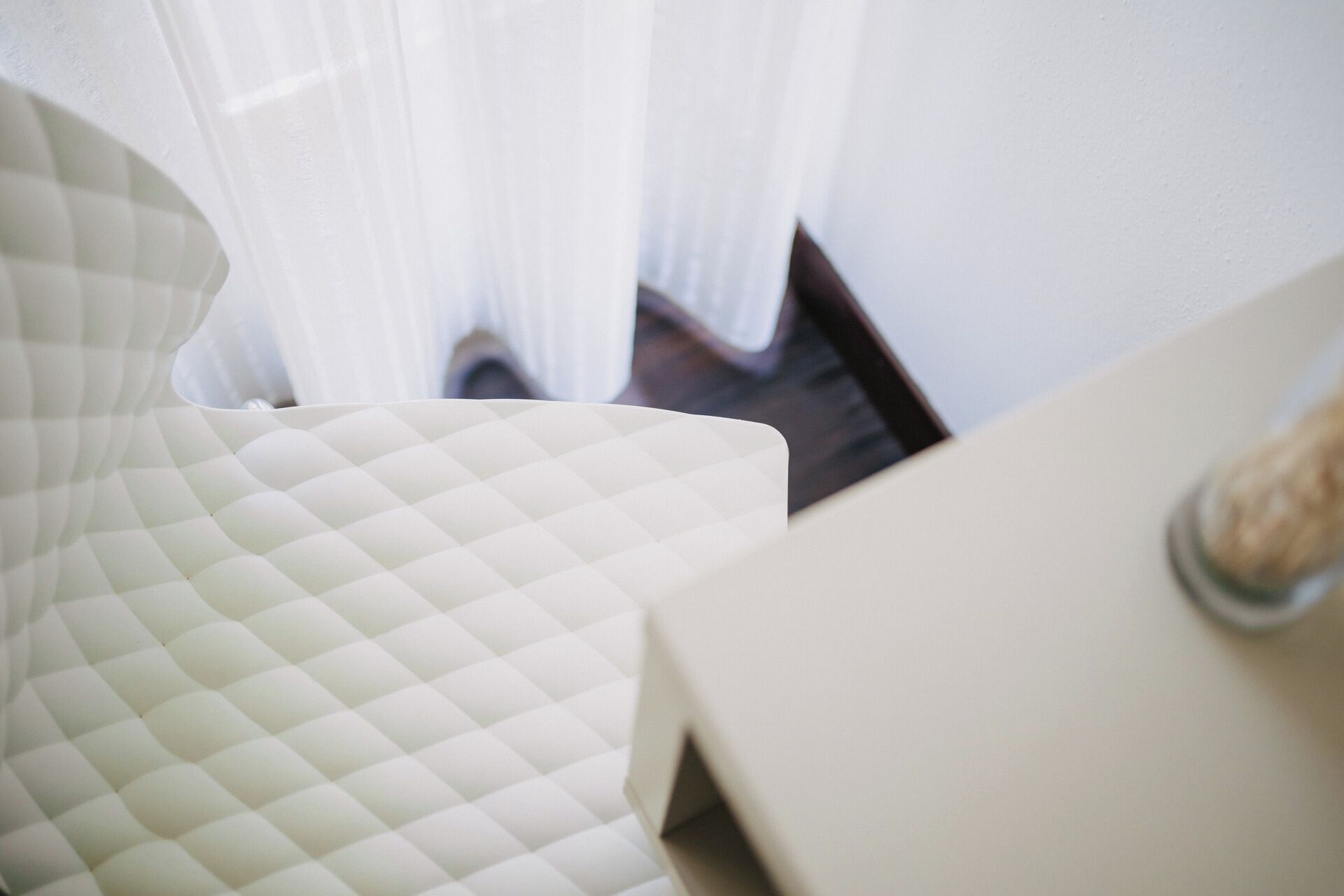 Una habitación con una butaca blanca acolchada y una mesa blanca con un objeto de vidrio encima.