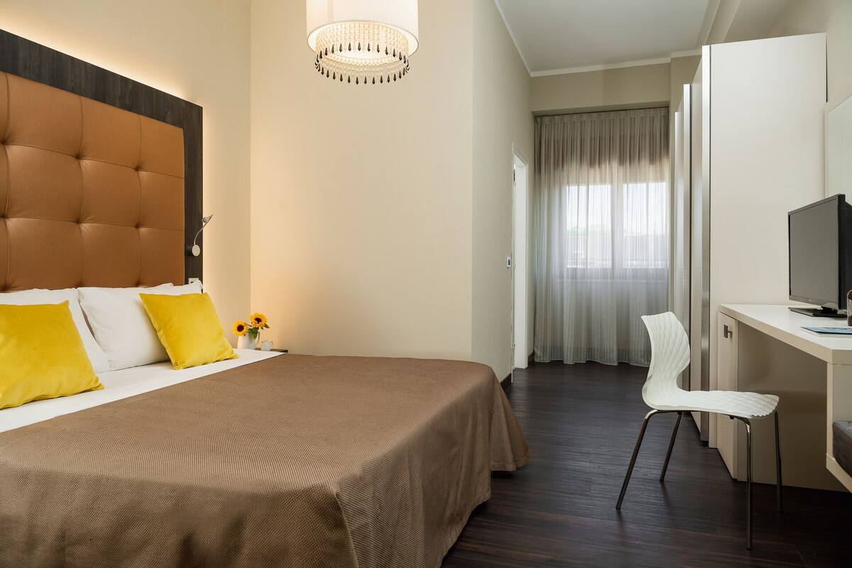 A hotel room with a large bed, a desk with a chair, a television, and a chandelier.