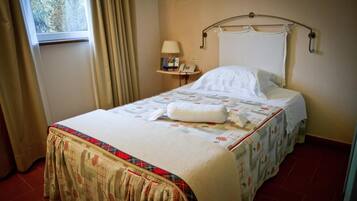 A neatly made bed with a floral bedspread and a matching skirt, a bedside table with a lamp and books, and a window with curtains.