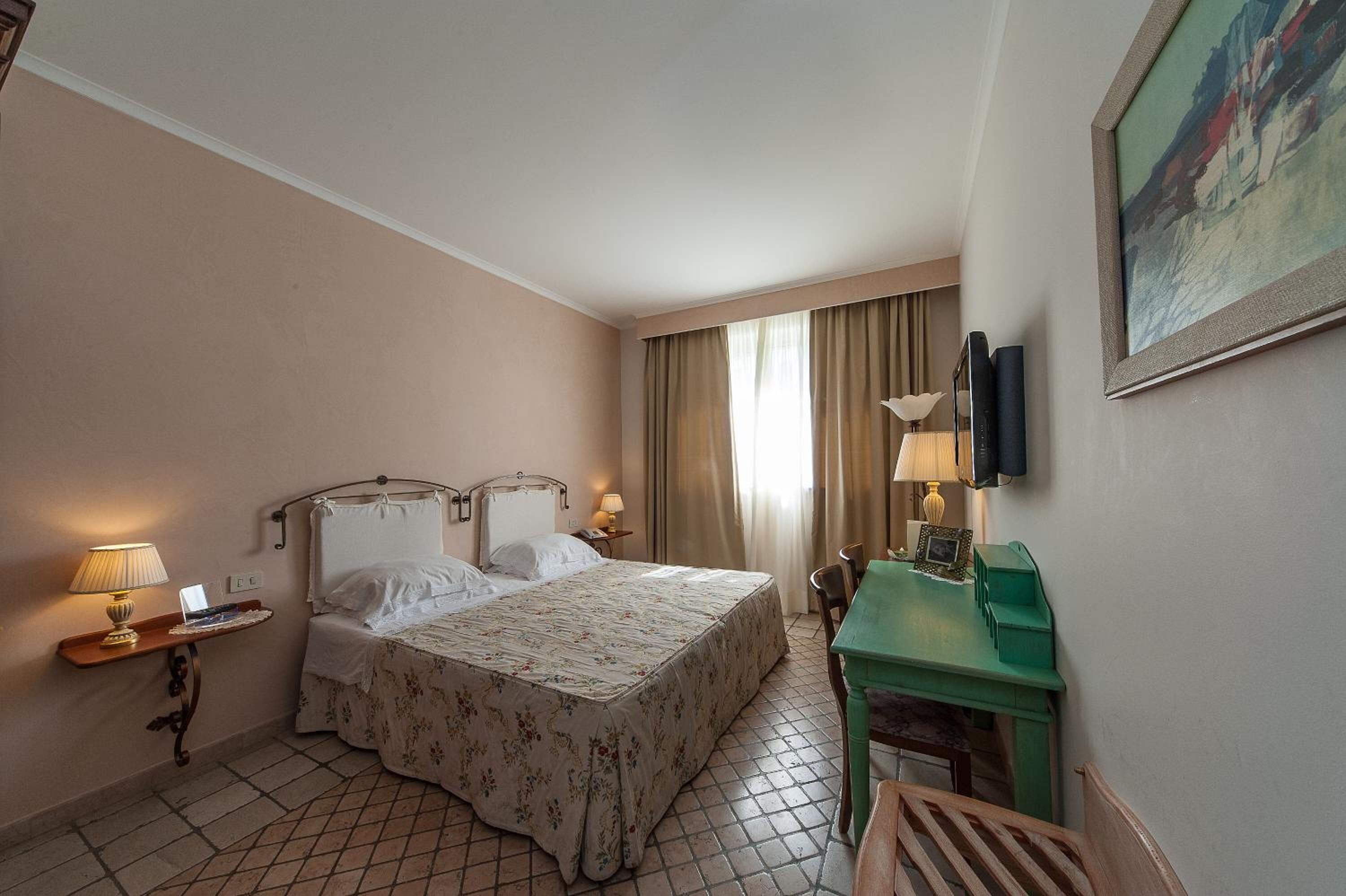 A hotel room with a bed, bedside lamps, a desk with a lamp, a chair, and a painting on the wall.