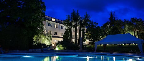 A swimming pool with a large building in the background, surrounded by trees and a covered area.