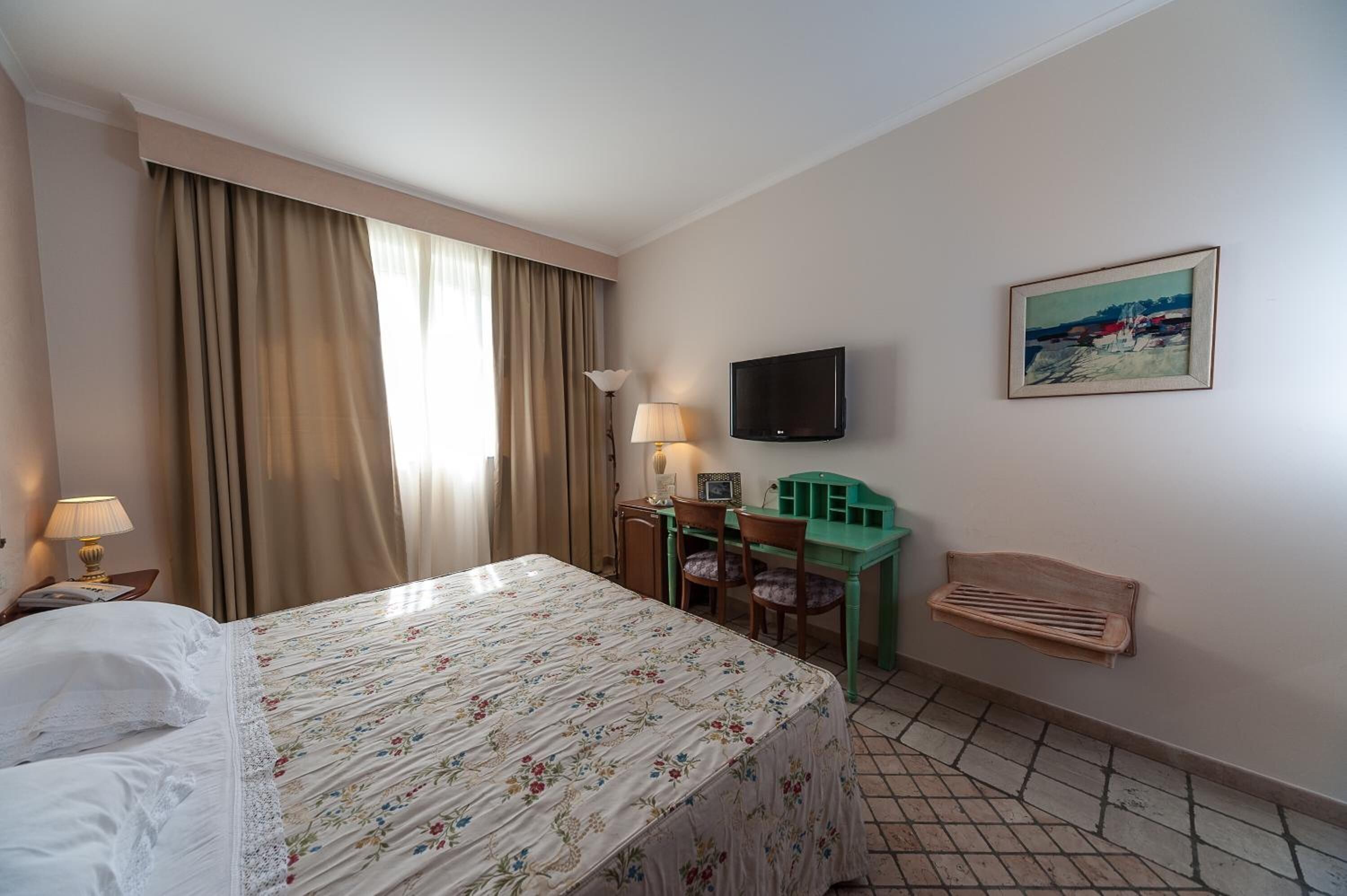 A hotel room with a bed, a desk with a chair, a television, and a painting on the wall.