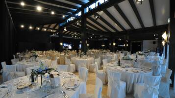 Une salle de banquet avec des tables rondes dressées pour un événement formel, agrémentée de nappes et de chaises blanches, et dotée d’un plafond haut avec des poutres apparentes.